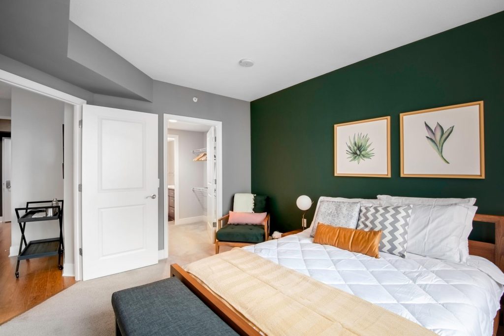 A furnished bedroom with a dark green accent wall and a large wooden queen size bed. The bed has lots of pillows as well as a few throw decorative ones and a yellow blanket. There is a grey chest for blankets at the end of the bed. The main door and the closet door are both open.