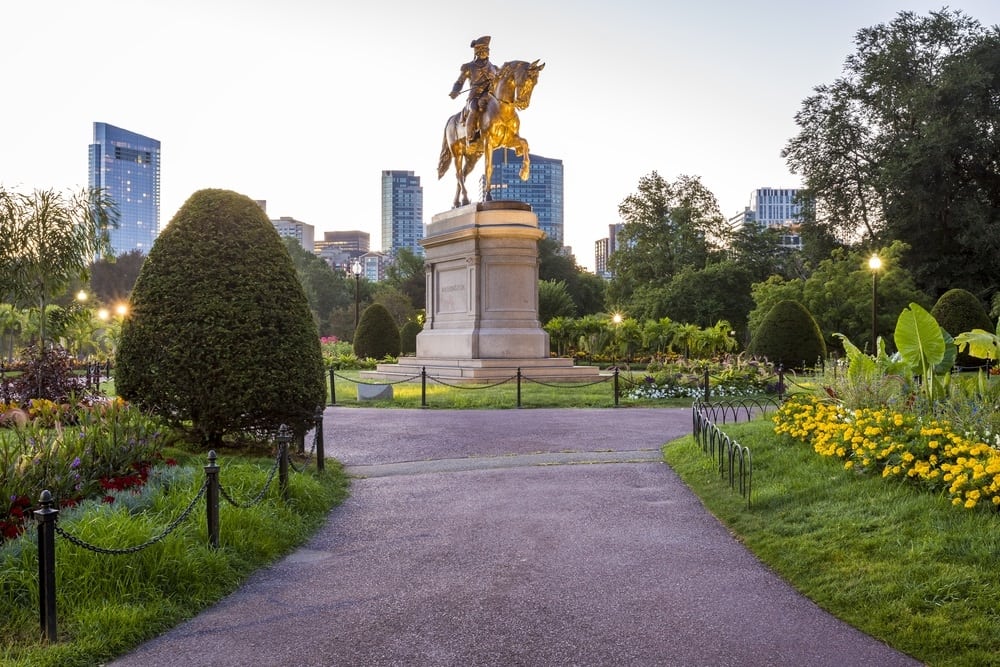 A gold statue of a man on a horse in the center of a park in Back Bay with greenery and the city skyline in the back. The sky above is grey