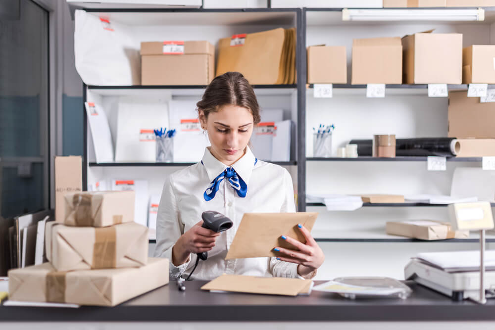 a woman working at the post office scans a package to find out the correct address before mailing it out. behind her are shelves full of letters and packages being sorted