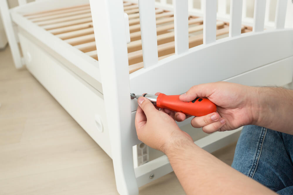 A hand is reaching out with a red screwdriver to take apart a white wooden crib in order to get it ready for a move