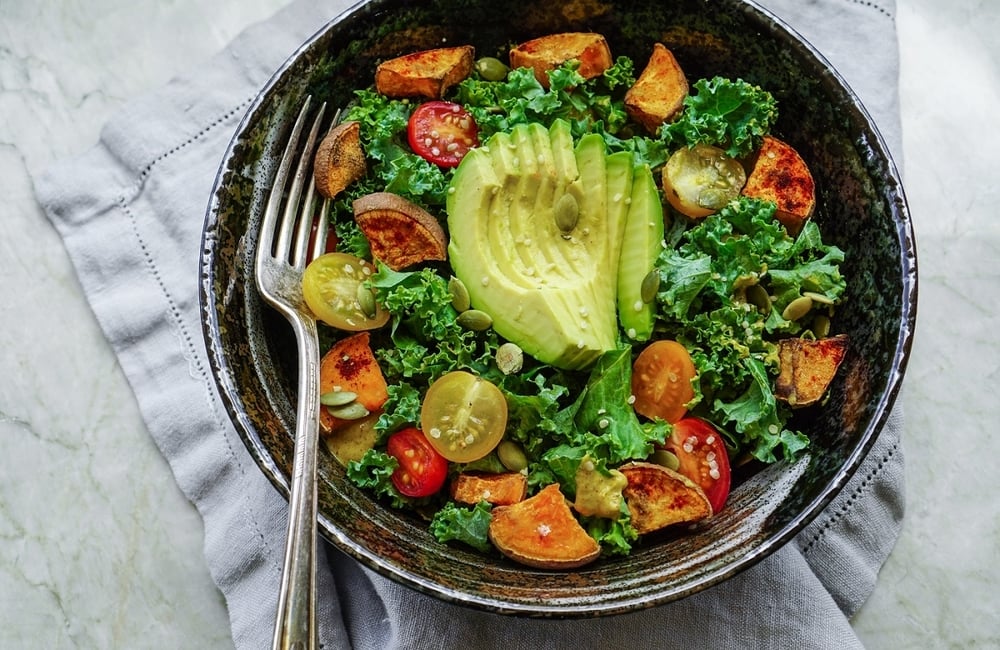 A view of a salad from above with greens and tomatoes and slices of avocado. There is a metal fork inside the bowl and it sits on a grey cloth, which is on top of a marble counter.