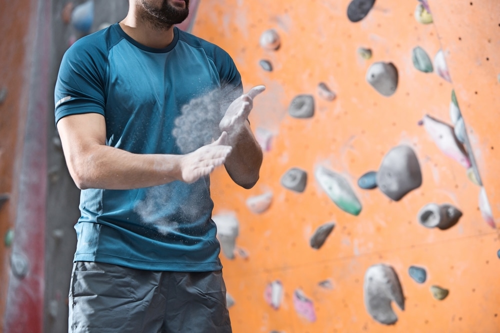 A man wearing a blue shirt and grey pants is standing in front of a rock wall. He is clapping his hands together to brush off some of the climbing powder. 