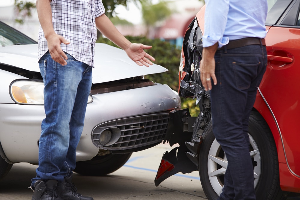 A man in blue jeans and a plaid shirt stands next to a silver car with an open hood and he is facing a man with dark blue jeans and blue shirt who is standing next to a red car with a broken back end. They are arguing after getting into an accident.