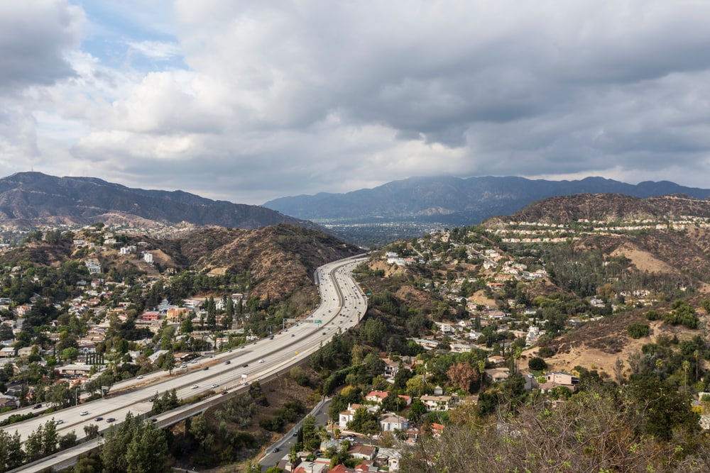 A view of Glendale from above, with a large highway passing through lush green mountains that are scattered with residential homes and buildings
