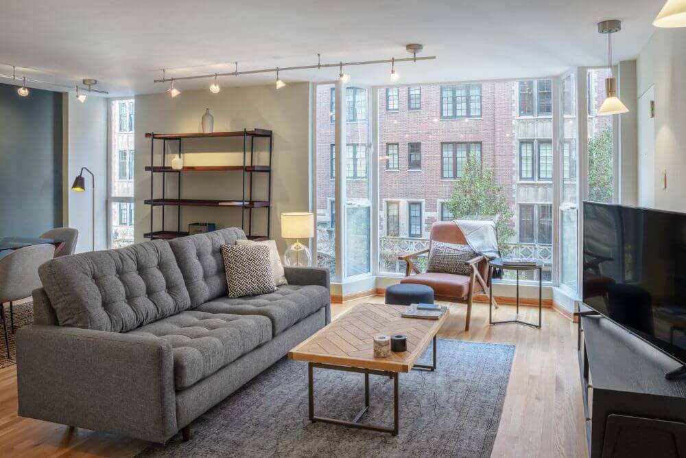 A spacious and furnished apartment in Chicago. There is a grey couch and a large glass bay window with a chair in front. There is an almost empty black bookshelf against the wall