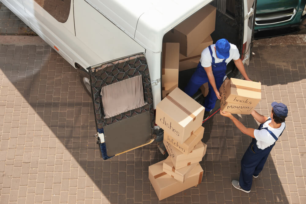 Two men wearing blue and white work uniforms are unloading boxes from a white moving van. The boxes have the designated rooms that they belong with written on them. This photo is taken from above