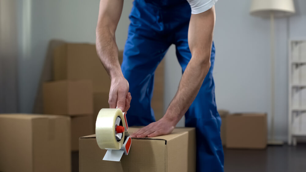 A man wearing a white and blue work uniform opens some packing tape has he closes a cardboard box