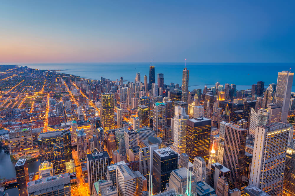 A view of the city of Chicago by night. There are lots of tall buildings all lit up and the streets are lit from the cars buzzing by. There is a view of the water as well that lines the coast