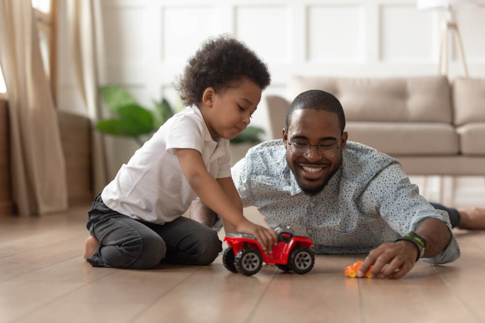 A man on the floor wearing glasses and a buttoned up shirt is smiling while playing with his child on the floor with toy cars. There is a small child next to the father wearing a white shirt and pushing a red car.