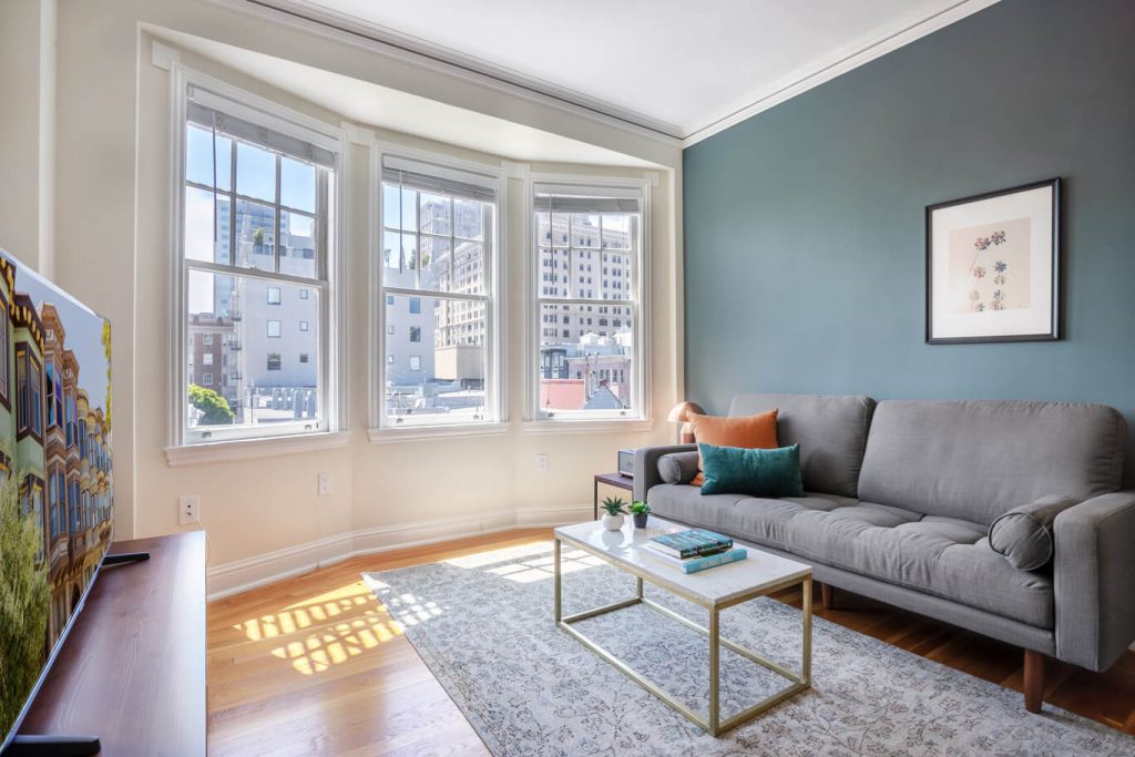 a minimalist apartment in san francisco with a large grey couch next to a large bay window. In front of the couch is a small marble and gold coffee table. There is a purple and grey carpet on the floor.
