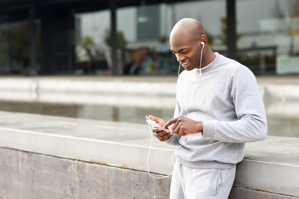 man standing in grey sweatshirt listening to audio on his headphones and smartphone