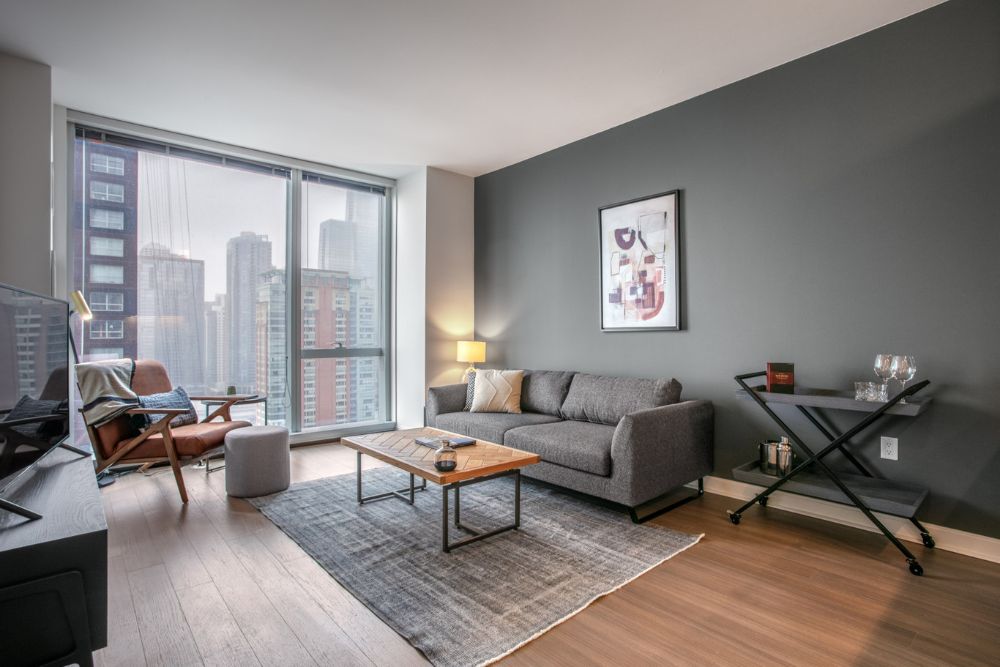 A furnished apartment with an olive green accent wall and a grey couch and a wooden coffee table with a black bar cart next to the couch. There is a large window with a view of the tall buildings of Chicago. Across from the couch is a brown leather arm chair with a blanket and a pillow