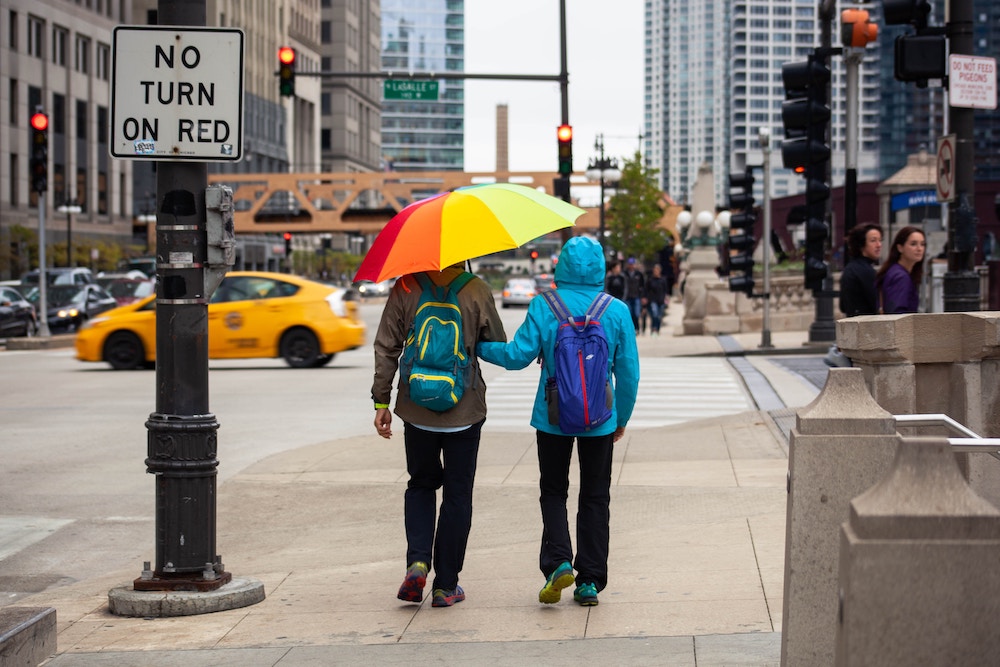Two people walking with an umbrella in Chicago. On person has a brown jacket and a blue and yellow backpack on while the other person has a blue jacket and a blue and red backpack. Their umbrella has rainbow stripes. A yellow taxi is driving past them.