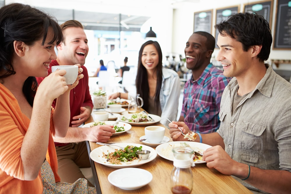 A group of five friends sitting around a table at a restaurant eating and drinking and smiling together. There are lots of plates and glasses on the table