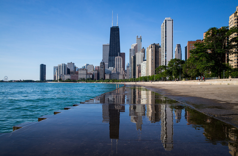 A view of many tall buildings in the Chicago skyline. With water on one side and pavement on the other and the buildings straight ahead.
