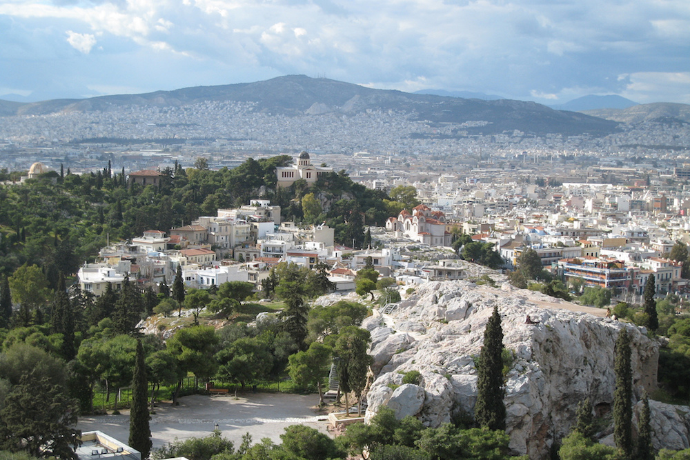 A view of the city of Athens and all it's buildings from above. There are many trees and a mountain straight ahead with a rock formation on the lower right side of the photo.