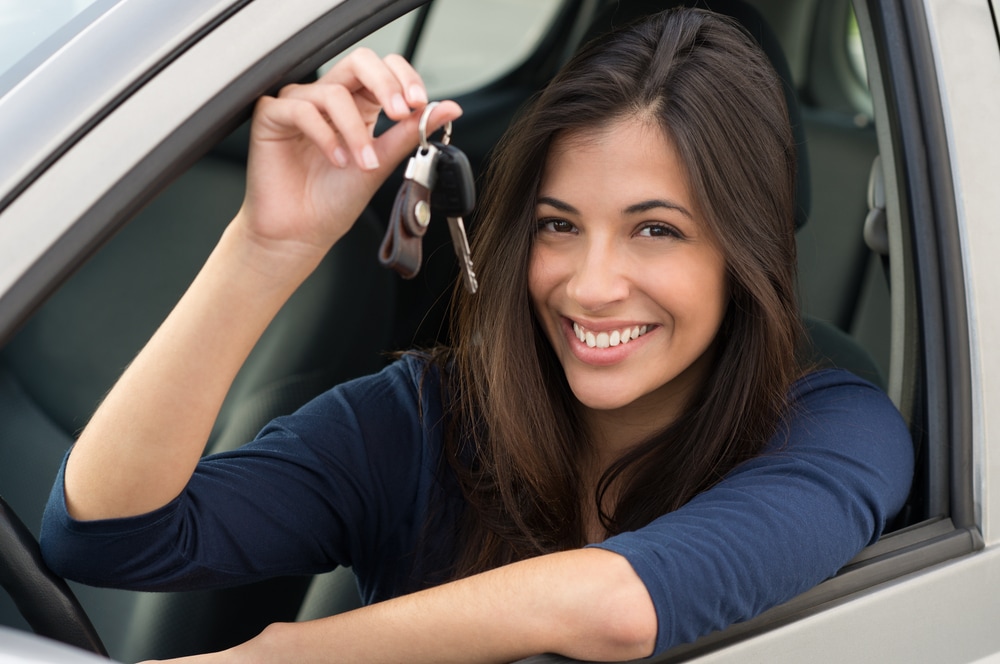 A woman with brown hair wearing a blue shirt sitting in the passenger side of her car holding up her keys and smiling