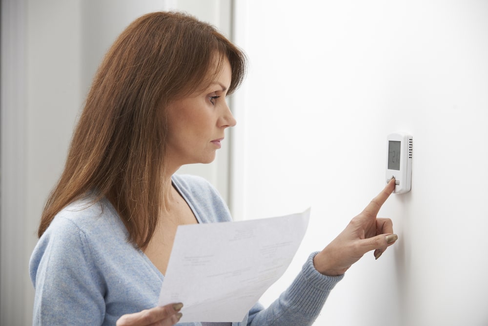 A woman with brown hair wearing a light blue sweater is holding a utility bill while adjusting the heating in her home.