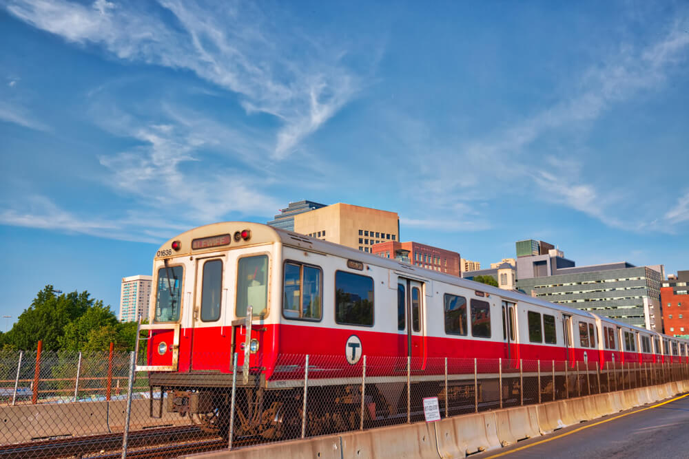 a red and white T train in Boston rides past some buildings and trees under a bright blue sky with wispy white clouds