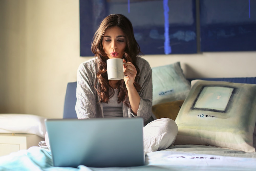 A woman is on her laptop while drinking coffee in bed