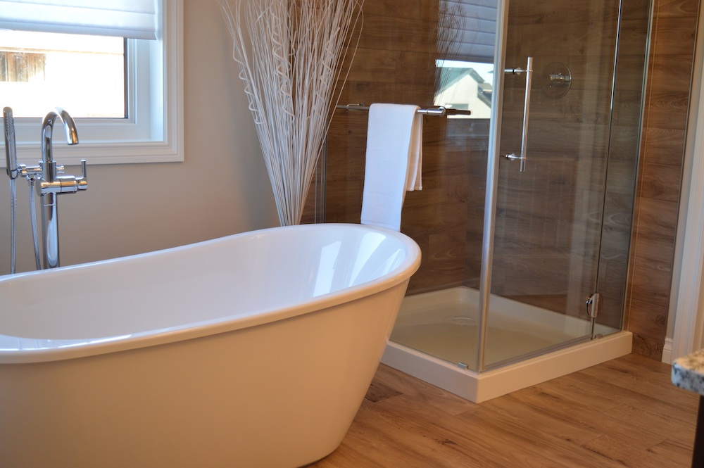 An upscale bathroom with a wooden floor and a wooden glass shower. There is a large white standing bathtub with silver details