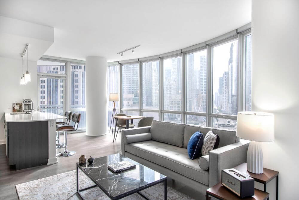 spacious and light fully furnished apartment in Chicago that is managed by Blueground. There is a large grey couch, a marble coffee table and behind that is a wall of windows
