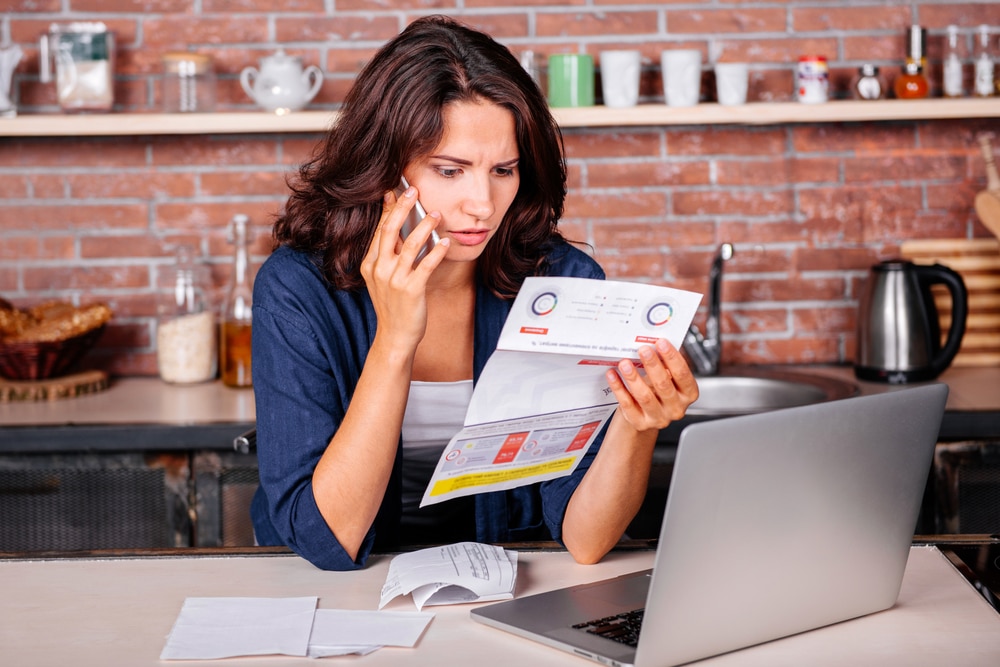 Young woman sitting in the kitchen with dissatisfied face in front of laptop holding utility bills, talking on mobile phone