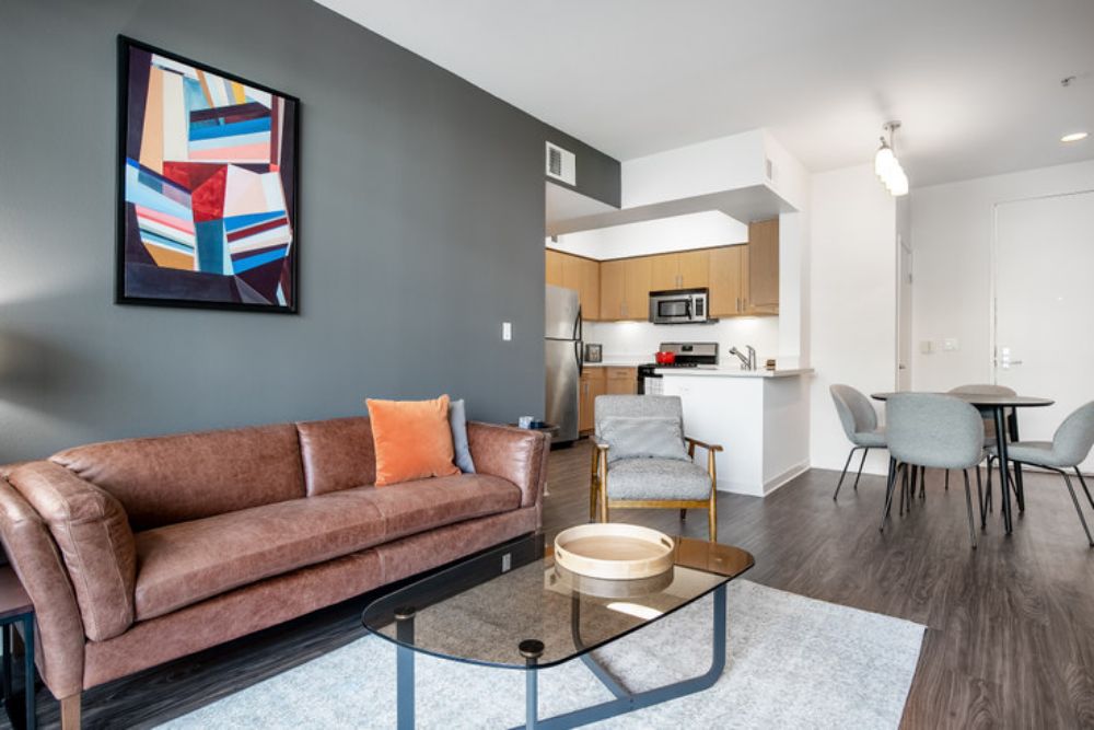a Blueground apartment in Los Angeles with an olive green accent wall and brown leather couch with an orange throw pillow. To the left of the couch is a small dining table with grey chairs while in front of the couch is a glass coffee table on a grey rug.