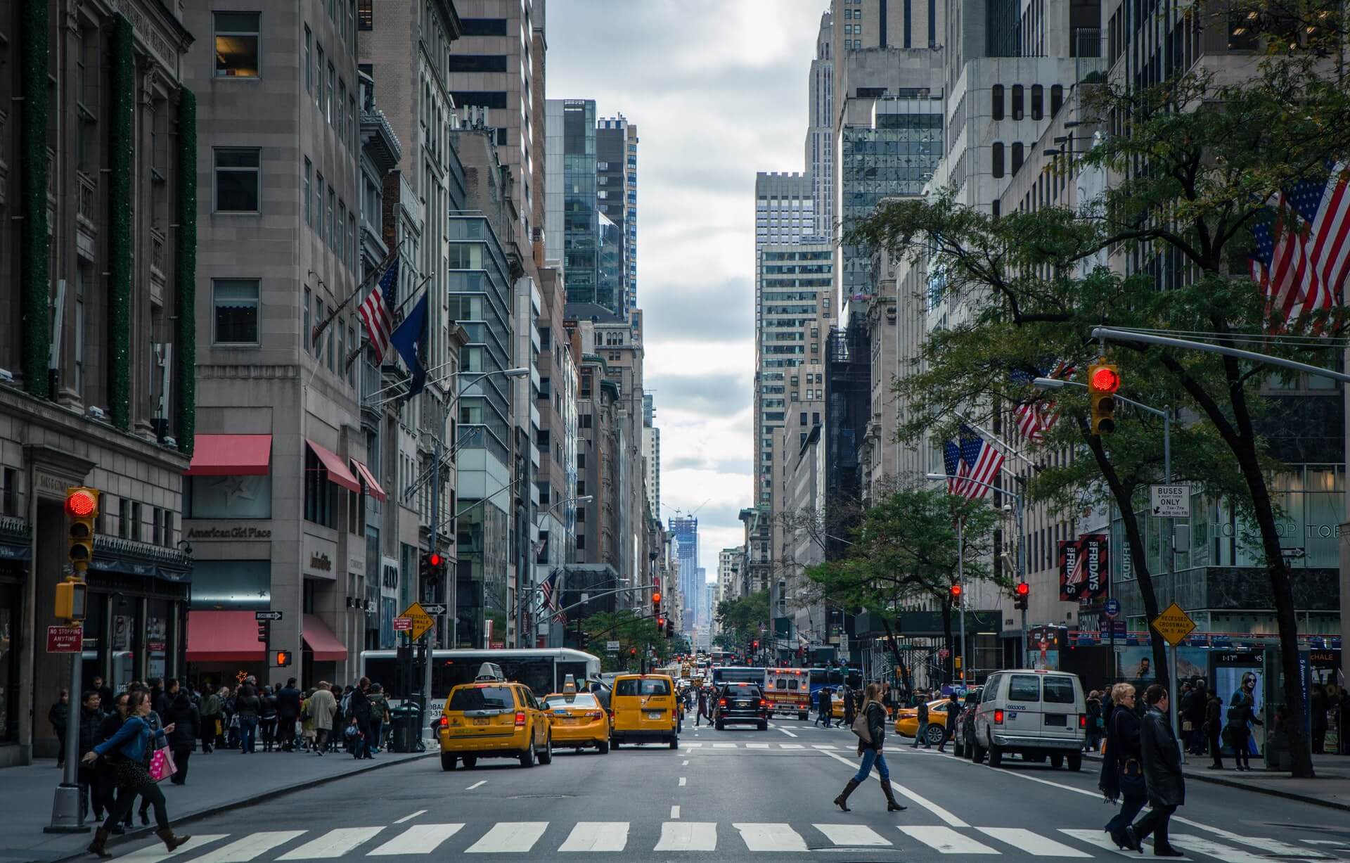 A view of a busy and hectic main street in New York City. There are lots of people walking around in all directions , there are lots of yellow taxis and cars around the road.