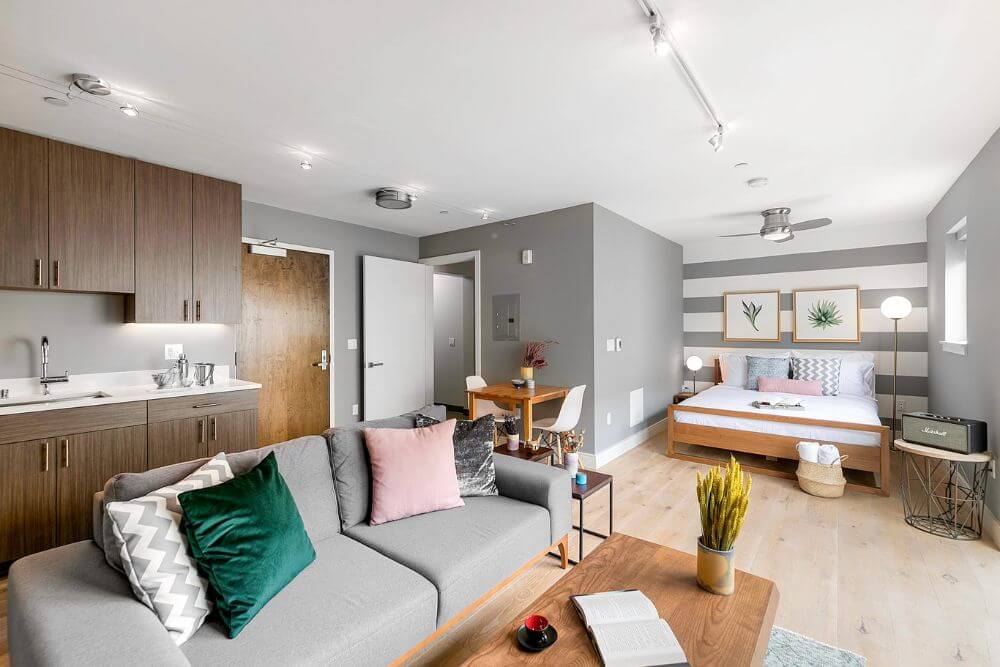 A fully-furnished and equipped studio apartment located in the 246 Ritch building in South Park San Francisco. There is a grey couch with green and pink pillows, in front of a kitchen with dark wood cupboards. The couch is sitting across from a large queen size bed.
