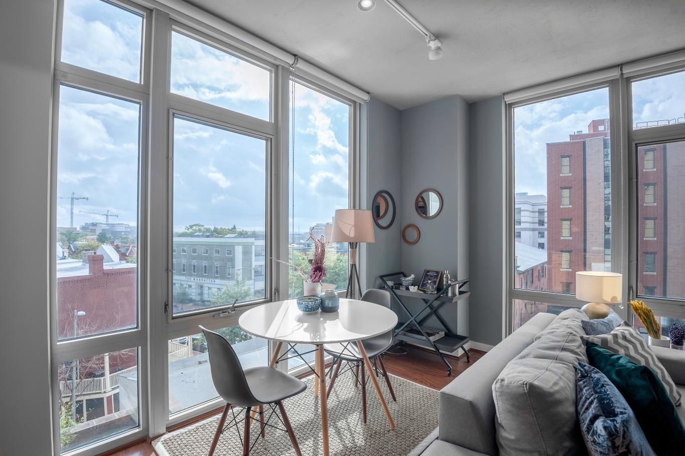 There is a white table and two grey chairs near a window in a dining area in a Blueground apartment in Washington, D.C. There is also a grey sofa with some green pillows on it.