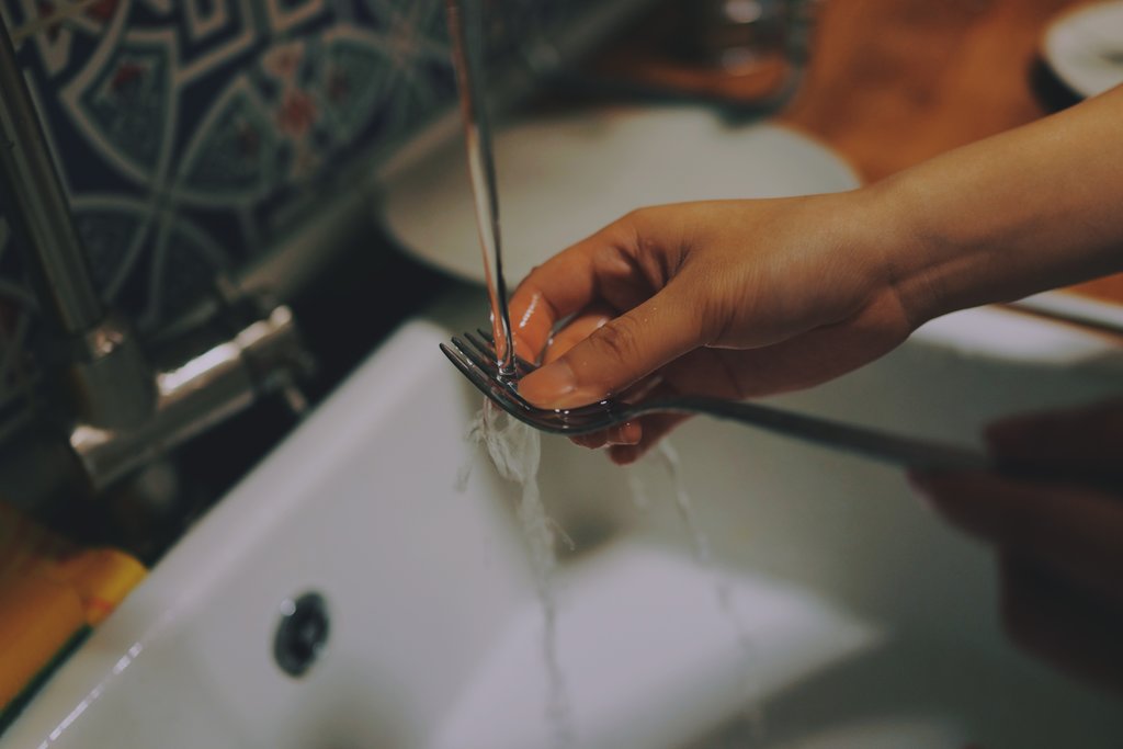 A woman's hand is extended out holding a fork and rinsing it off under the sink faucet. The sink faucet is switched on, the water is running and it runs on the tongs of the fork over a large, deep white sink. A bit of the vibrant tile back splash is visible behind the sink.