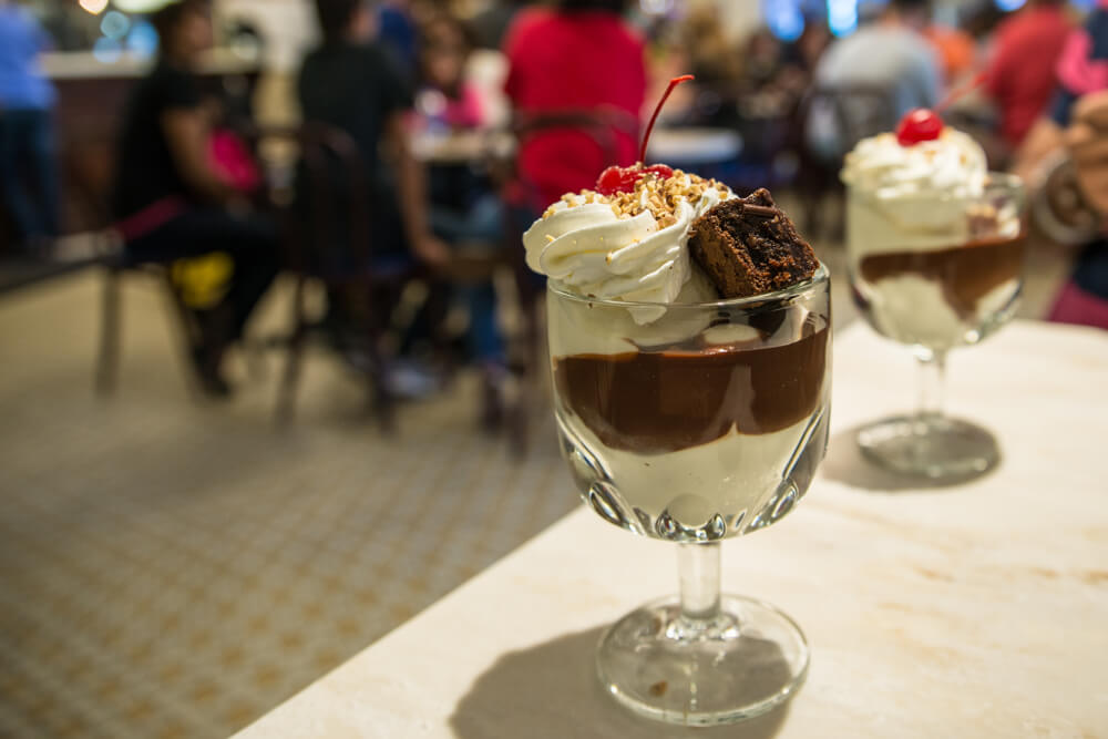 two world famous chocolate sundae bowls on a white table at Ghirardelli in San Francisco. There are many people sitting around a table in the back eating their desserts.
