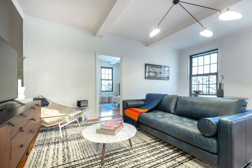 A furnished apartment in New York City with a dark grey leather couch and a small white round table and in the corner there is an armchair. There is a blanket tossed onto the couch and another one tossed onto the armchair. Above the leather chair is a large black light fixture