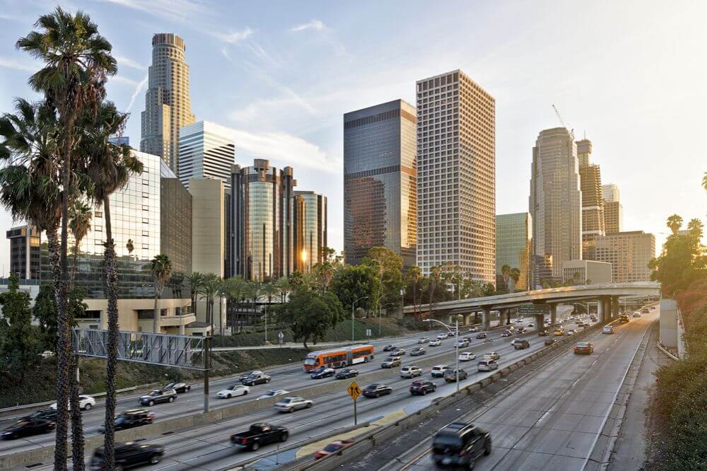 A view of some buildings in downtown Los Angeles during sunset In front of the buildings is a highway with a lot of cars driving both ways