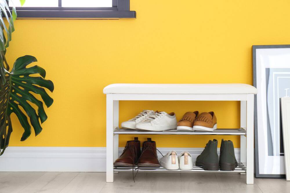 A small white shoe rack with two shelves full of shoes sits against a bright yellow wall under a framed window. To the left of the shoe rack is a large, leafy green plant.