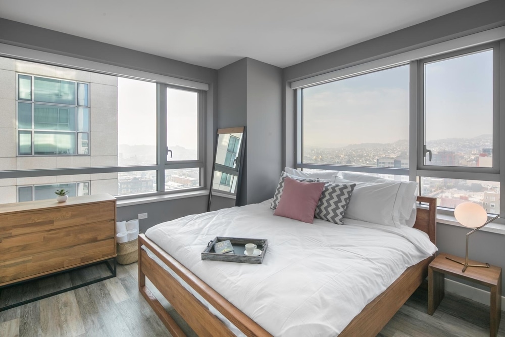 a bedroom with a lot of windows that are letting in natural light and offering a view of the city. There is a wooden bed and dresser and nightstand with a lamp on top