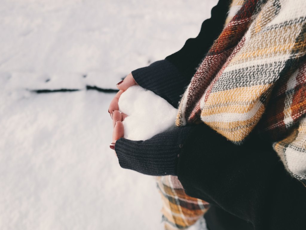 A woman wearing a black sweater and a plaid yellow and red scarf is standing outside in the snow and making her hands into a heart shape. In her hands she is holding some snow, creating a heart shape with the snow. 