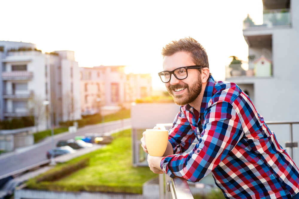 man standing on a balcony holding a yellow coffee mug and smiling and staring off into the distance