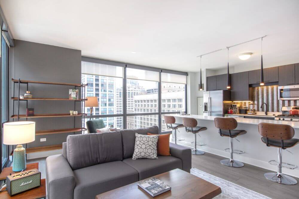 a furnished apartment in Chicago with a grey couch and a wooden coffee table with a book on it. There is a wall of windows next to the long white kitchen island. There is a wooden shelf with some decorative items on it next to the window. Next to the couch is a Bluetooth speaker and a blue lamp with a white lamp shade. The white kitchen island has black and brown bar stools.