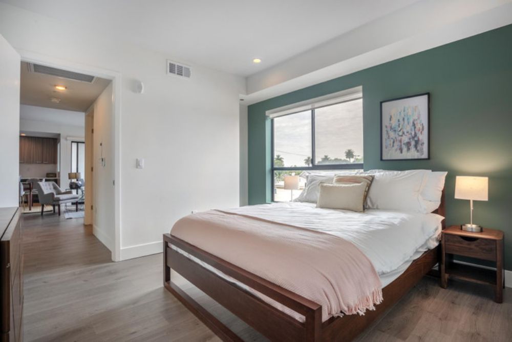 a Blueground apartment in Los Angeles with an olive green accent wall a large white bed in a wooden frame and a small wooden nightstand with a lamp on top next to the bed
