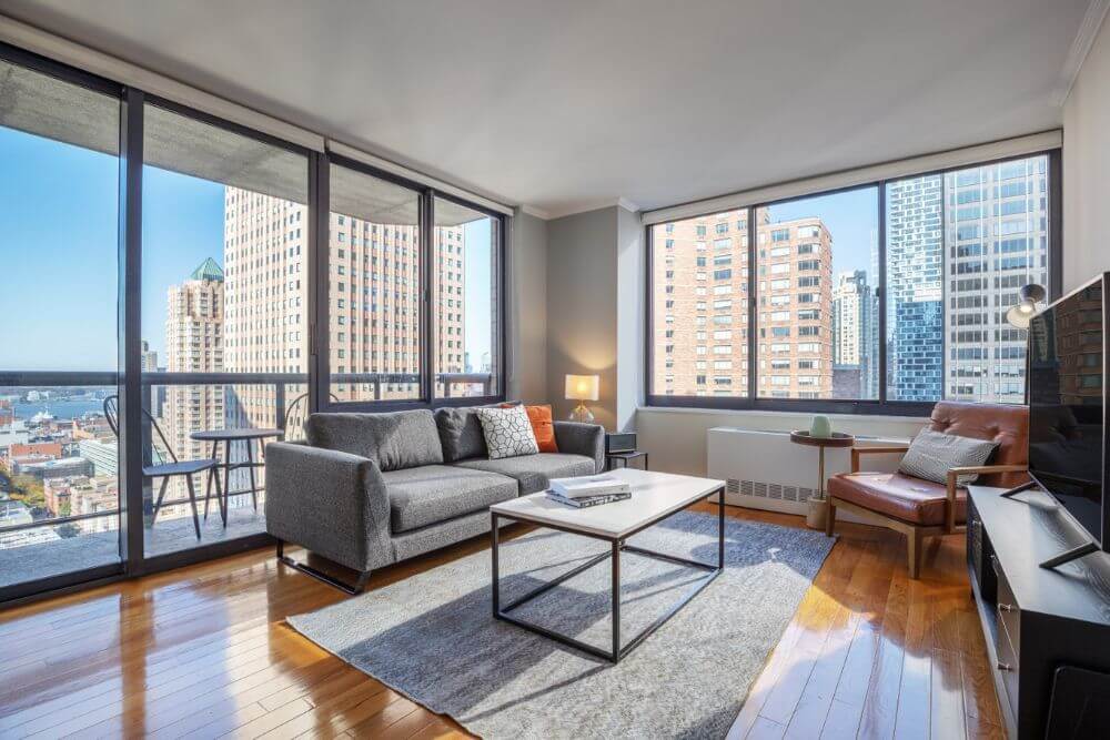 An apartment in New York City with two walls full of windows and a grey couch and a coffee table and TV stand. There is a set of table and chairs on the balcony.