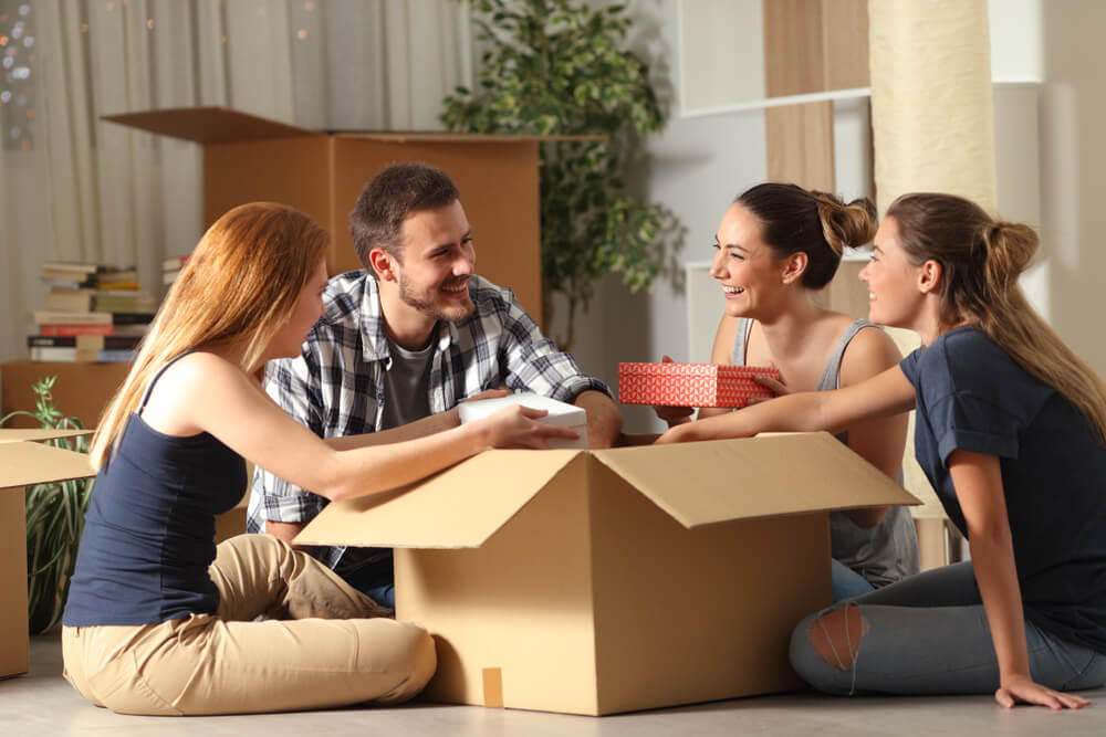Three women and one man are sitting around a large cardboard box in the middle of the living room laughing and talking together