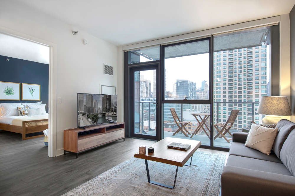 a living room with a dark grey couch, a wooden coffee table and a TV next to a small balcony that provides a view of the buildings of Chicago