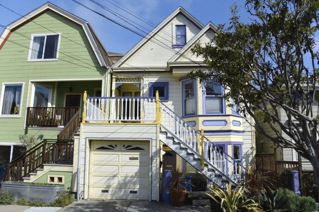 A blue and white Victorian home with yellow trim and a garage in San Francisco. There is a green house attached on the left and a smaller grey house on the right. There are plants as well as a tall tree.