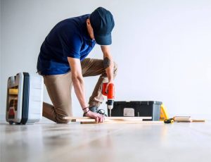 A man wearing a blue shirt and a blue hat and brown paints is working with pieces of wood and power tools