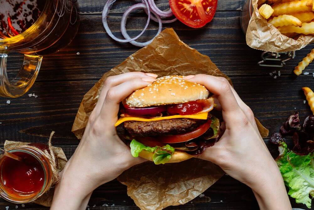 a person is holding a burger over a table that is filled with french fries and toppings