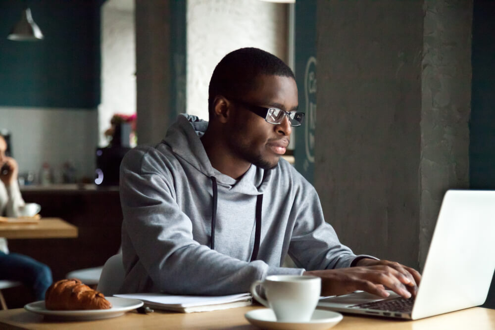 dark skinned man wearing glasses and a grey sweatshirt sits at a table in a cafe while working on his computer with a croissant and a coffee in front of him 