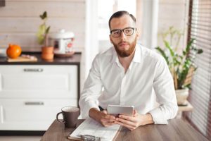 A man wearing a white shirt and black glasses and holding a tablet sitting at a kitchen table ready to sign a paper that is on a clipboard in front of him. Near his right elbow is a brown mug. Behind him are some white kitchen drawers with a brown countertop