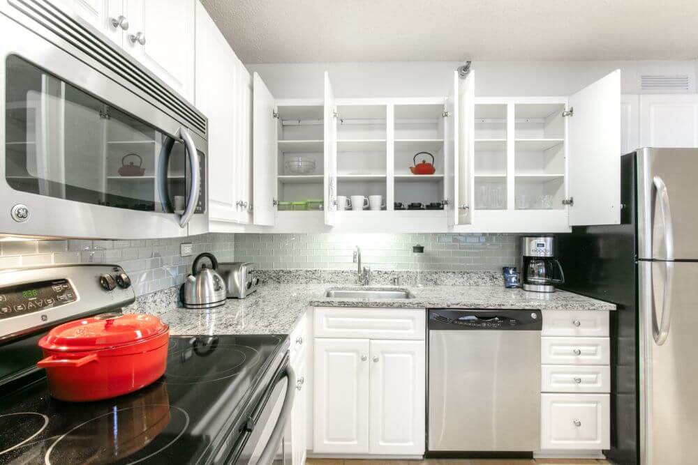 A fully equipped kitchen with white counter tops and stainless steel appliances. All of the cupboards are stocked with plates and cups